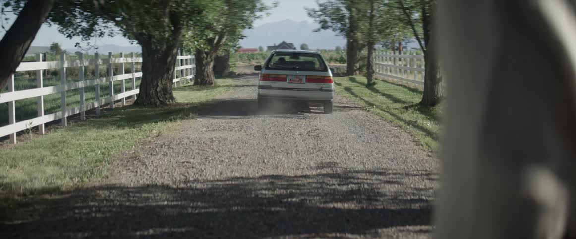 A person watches as a vehicles takes off down a gravel driveway