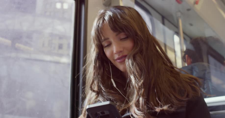 A woman sits on a city bus listening to something from her cell phone with ear buds