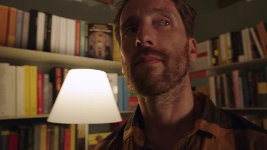 Camera moves towards man in dimly lit room surrounded by books