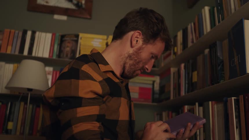 A man reads the back of a book and then puts it back on a shelf