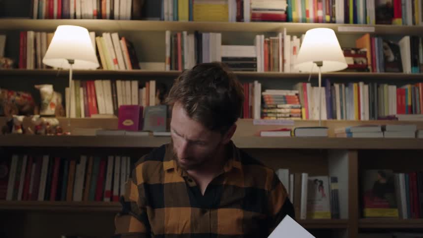 Man in plaid shirt looking through a book in front of bookshelf filled with books