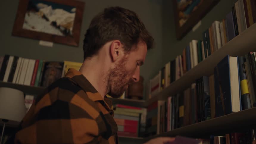Male chooses a book from the top of the book shelf and turns it over to read the back of the book