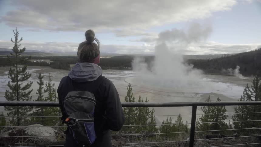 A woman stands at a railing overlooking steaming and smoking geysers in a park