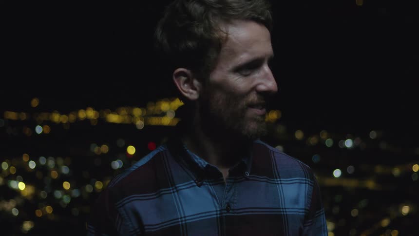 A bearded white man smiles with city lights visible behind him