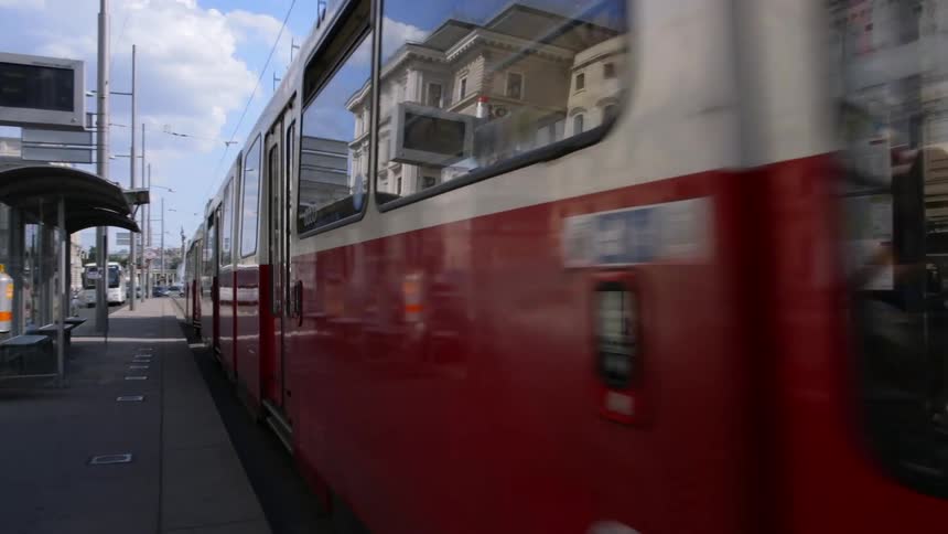 A trolley train travels on street level tracks past a bus stop on a sunny day