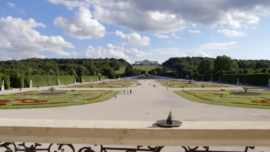 A balcony wall with wrought iron inserts overlooks a beautifully landscaped garden with sculptures along the hedges and people walking on paths