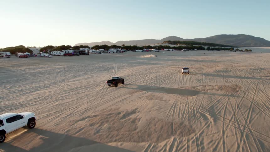 Trucks and motorcycles race across a desert in view of a row of recreation vehicles