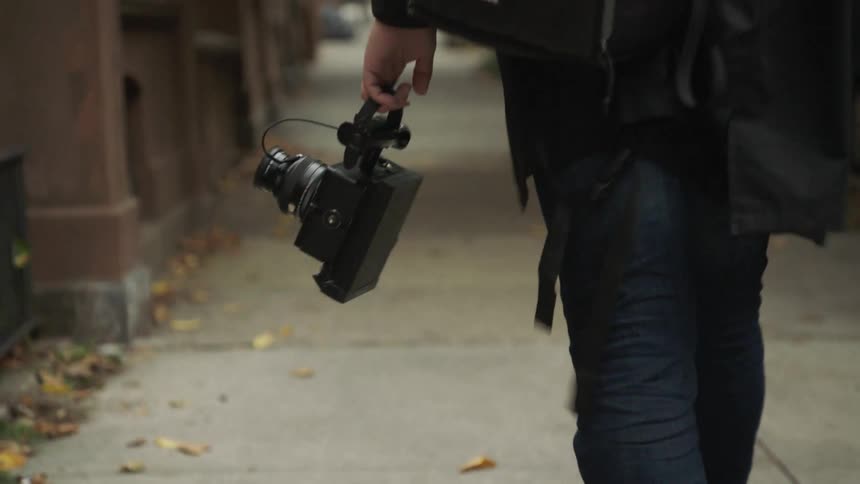 A white man carries a camera as he walks down a neighborhood sidewalk