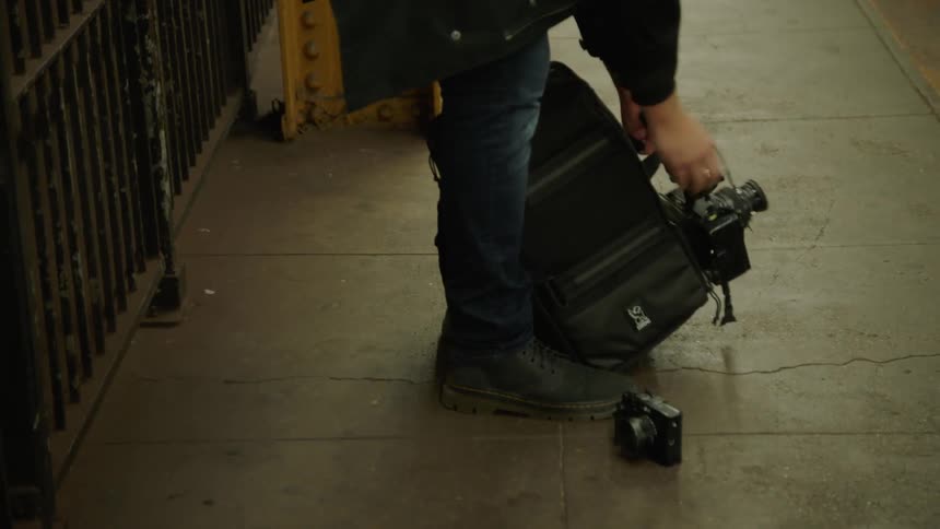A man stands on a city sidewalk and removes a camera from his backpack