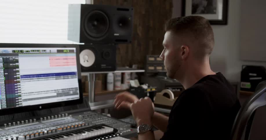 A white male musician studies a computer monitor while motioning with his hands