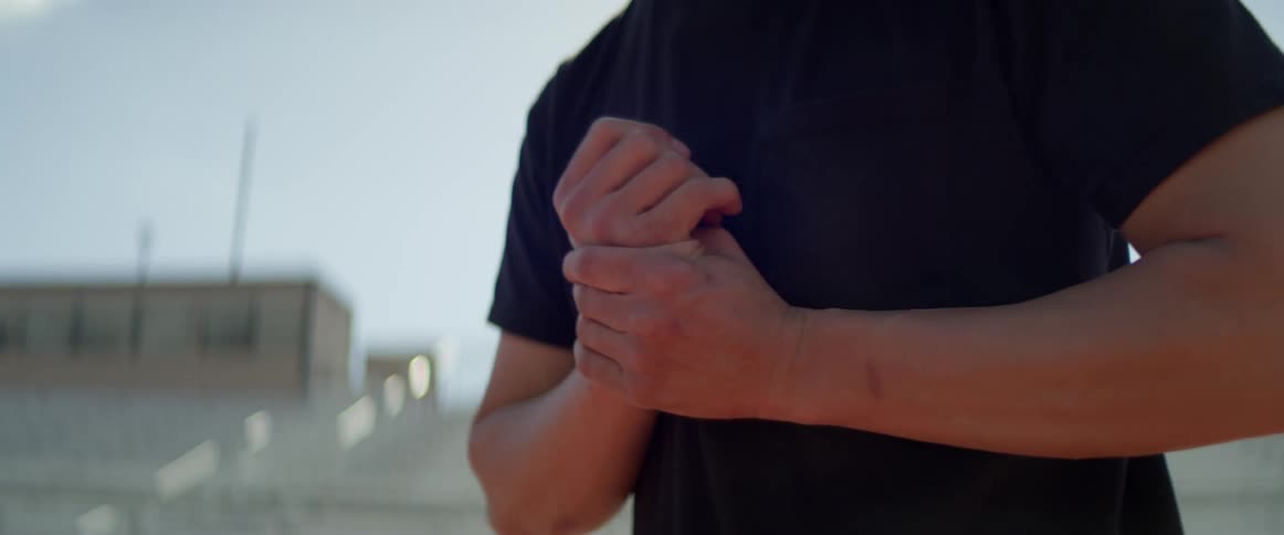 A man wearing a black tee shirt stands in an empty stadium rubbing his wrist on a bright sunny day