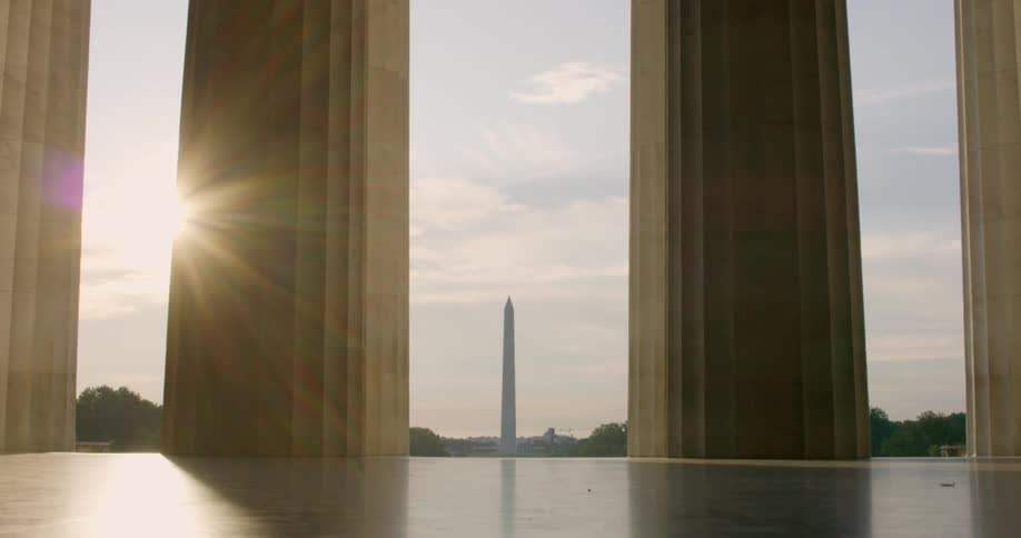 Sun shining brightly throught the columns of the lincoln memorial and the washington monument can be seen in the far distance