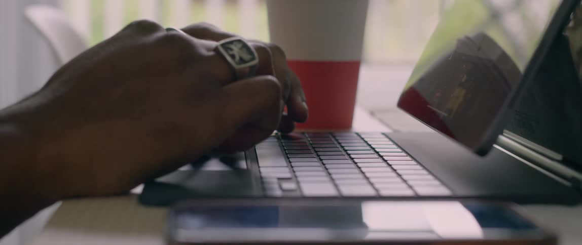 A person sits at a table scrolling through a laptop with a drink beside them