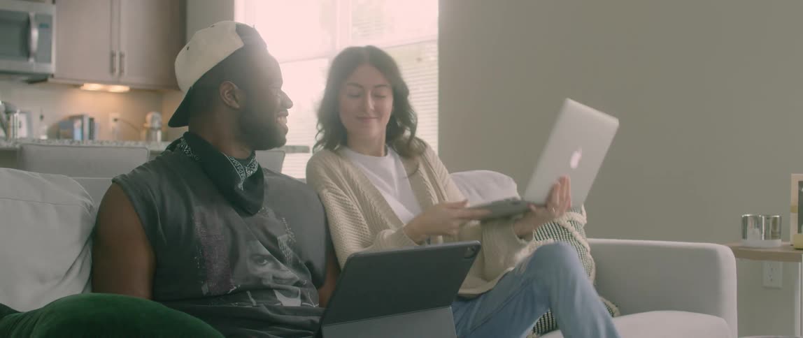 An asian woman walks over to a couch and sits down next to a black man