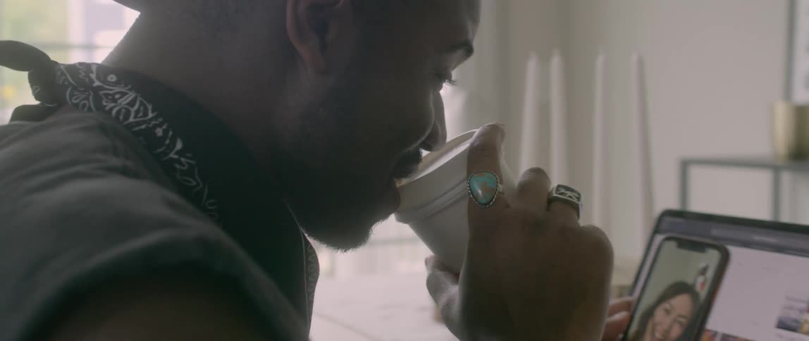 A black man drinks from a cup while looking at a laptop