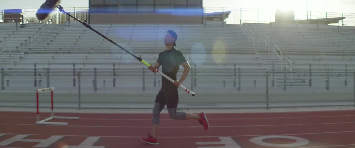 A man carries a mic on a long pole and follows a woman jogging on an outdoor track while others film her