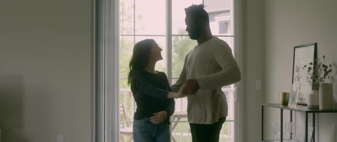 A black man and an asian woman dance in a living room