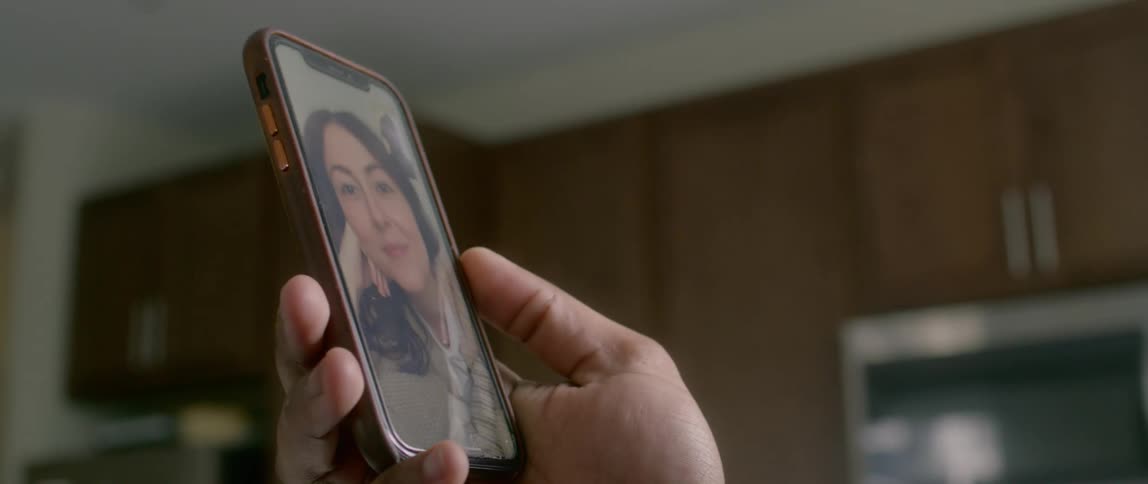 Male stands in front of the window in a kitchen holding a cell phone as he video chats with a female