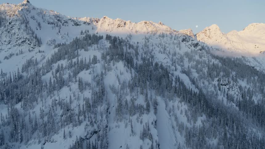 Tilt up reveal of snow covered mountain range with the moon between peaks