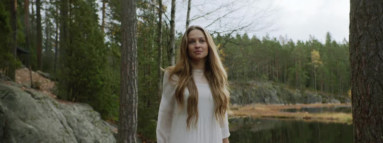 A white woman in a white dress walks along the shore of a lake