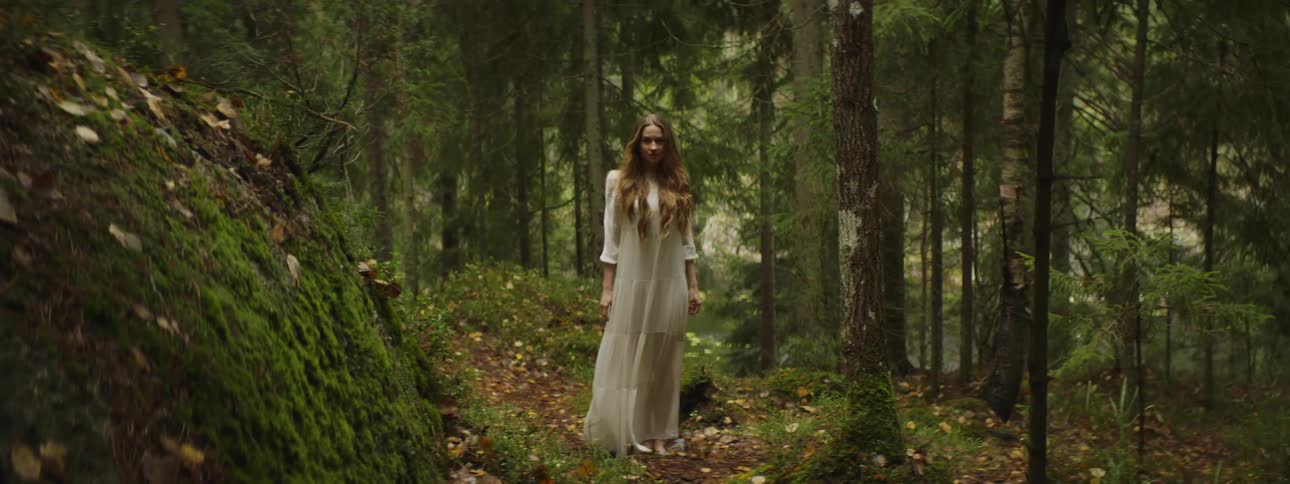 Woman in white flowing gown stands on path in dense forest