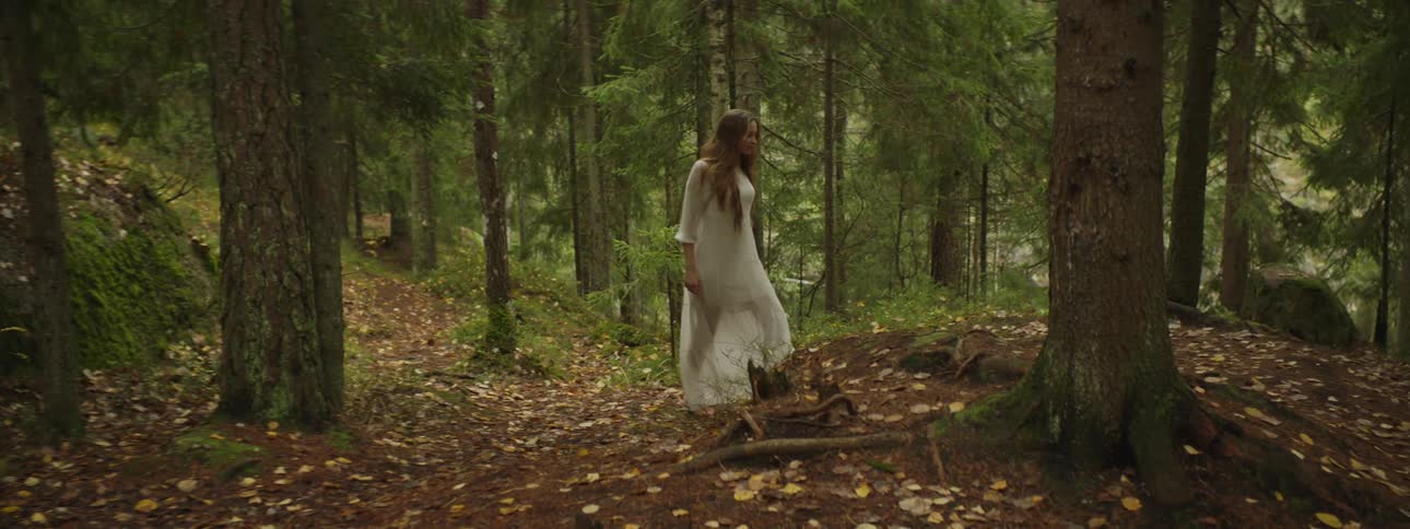 A woman wearing a long gauzy white dress walks barefoot in the woods
