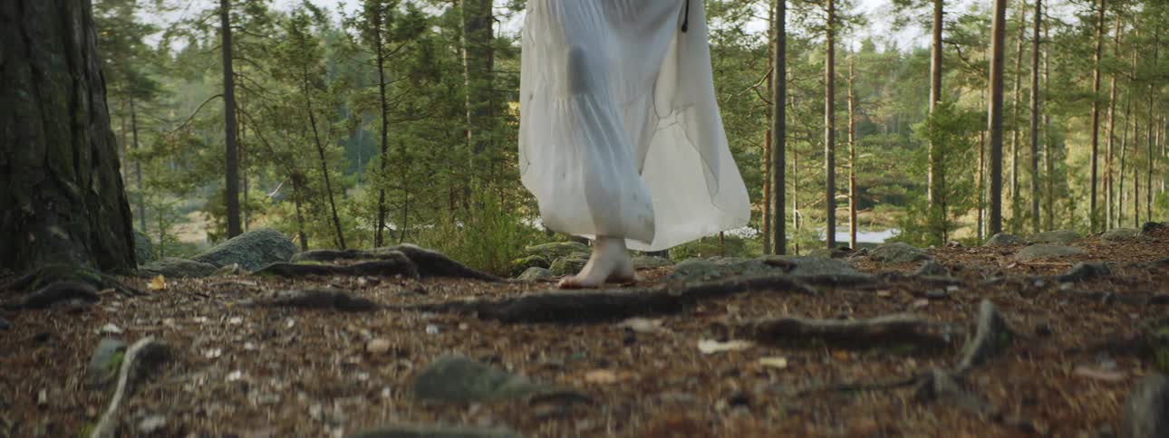 A woman lifting the bottom of her gown as she walks barefoot through a woodland