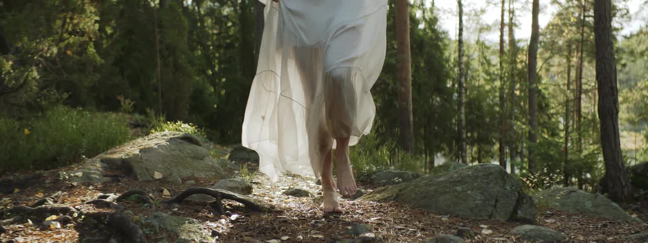 A woman in a sheer white dress walks barefoot in a forest