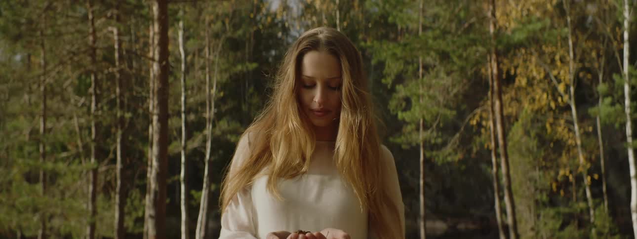 A white woman stands in a forest while holding an object in her hands