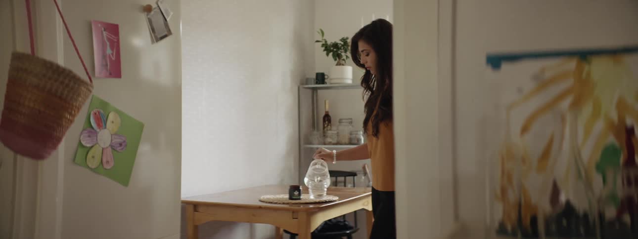 A woman stands at a table and pours hot water from a clear glass kettle into a clear cup on a tray