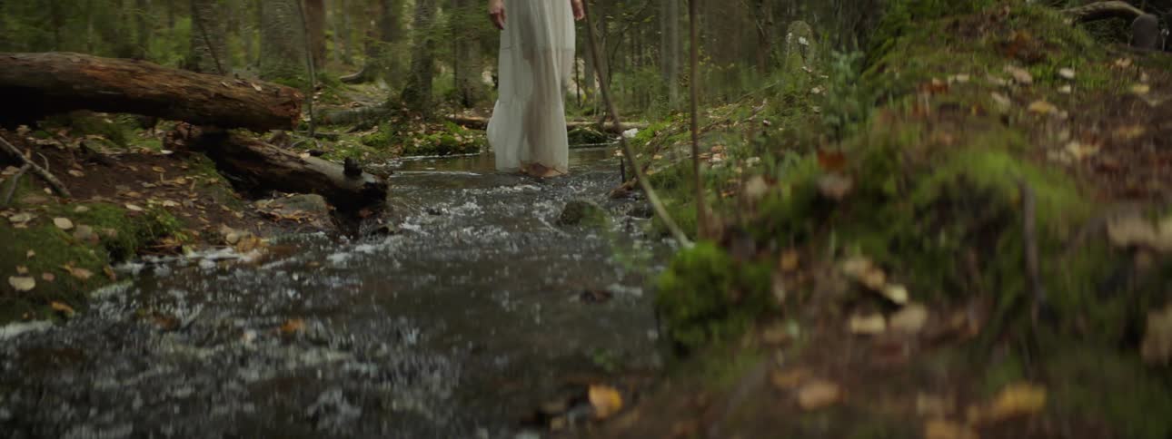 A white woman in a white dress walks across a shallow stream in the woods