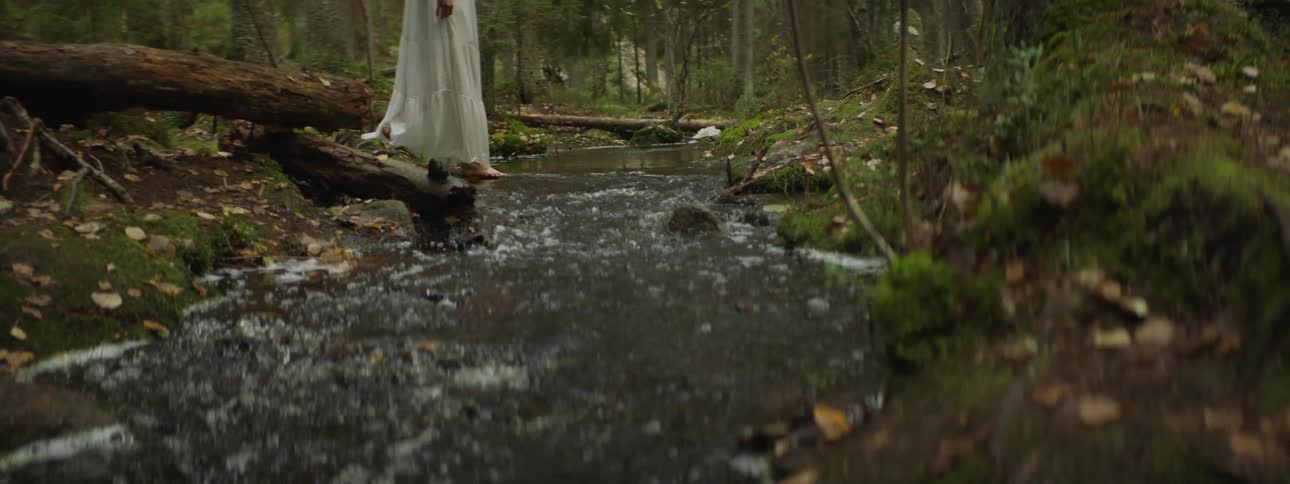 A woman in a white skirt crosses a stream barefoot