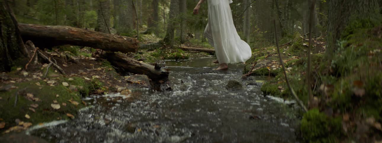 Female wearing long white sheer dress steps slowly into the water of a creek with her bare feet in the middle of the woods and begins to walk across the water as she tries to balance herself