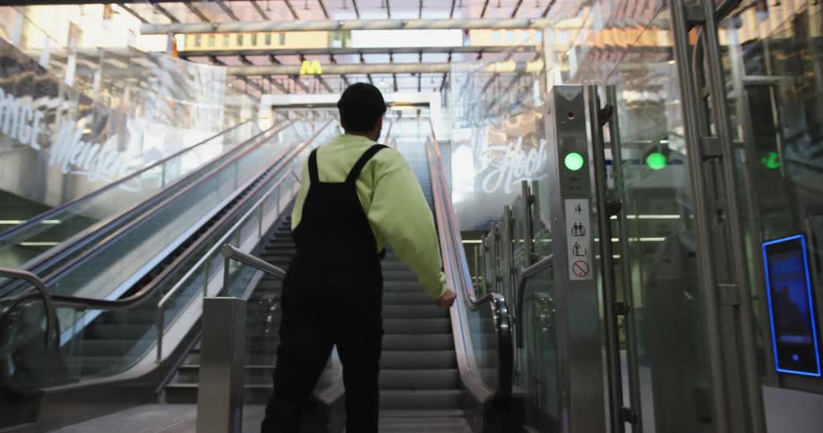 A white man in overalls walks and dances over to an escalator