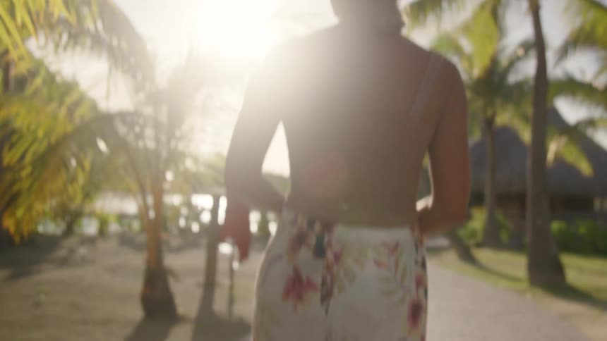 Woman in long flowing pants and tank top walks along stone pathway under palm trees
