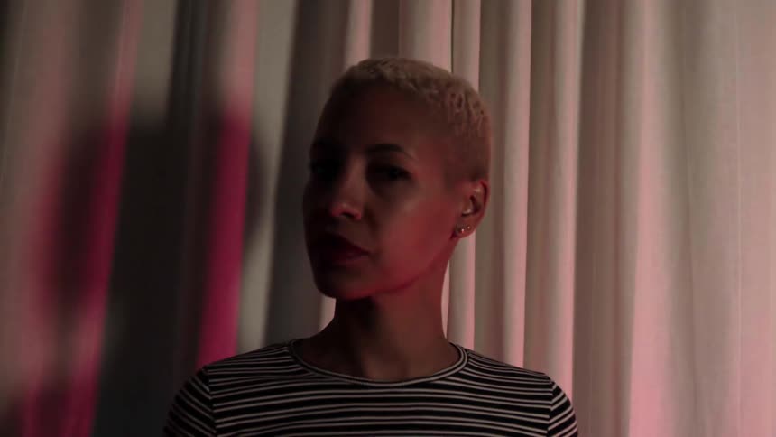 A black woman with short hair turning her head while keeping her eyes on the camera as the lighting in the room fades