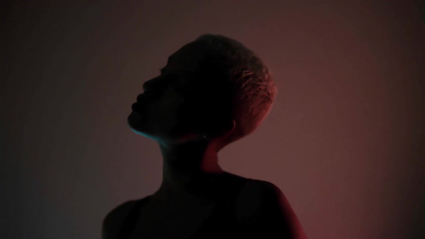 A shorthaired woman looking up at the ceiling and then down at the floor in a dark room with a dimly flashing colored light