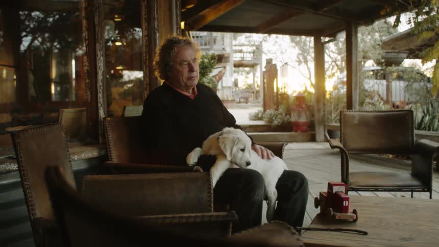 Male sitting on chair under roof of patio petting a golden retriever puppy that is sitting on his lap while the sun begins to rise