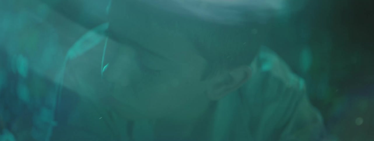 A boy lying on the ground outdoors at night turns his head as a bright green light begins to shine behind him