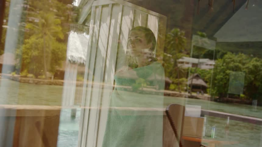 A woman with wet hair and wrapped in a towel glances out a glass window
