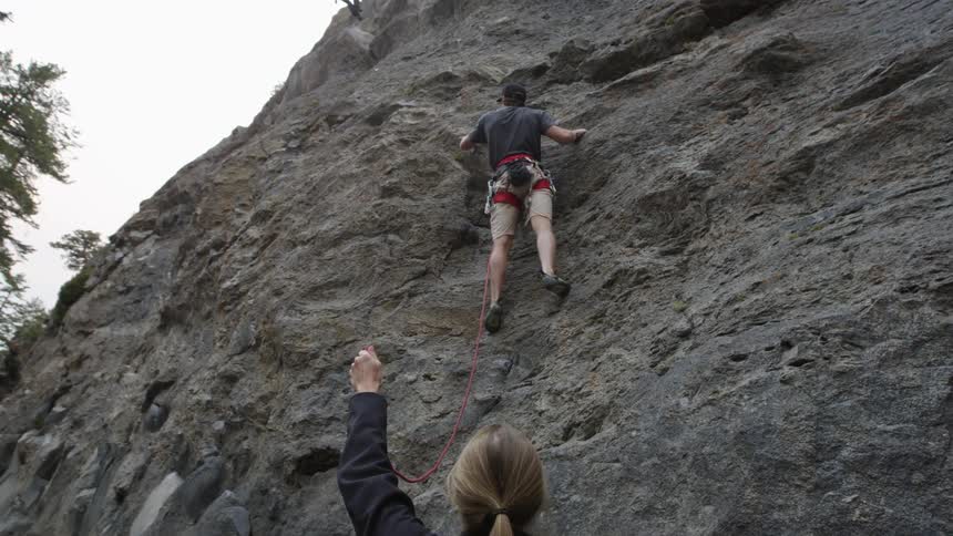 Male rock climber holds on to a rock while a woman below holds his rope