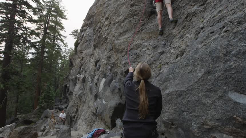 A man climbing a cliff with a woman standing at the bottom of the cliff giving him rope
