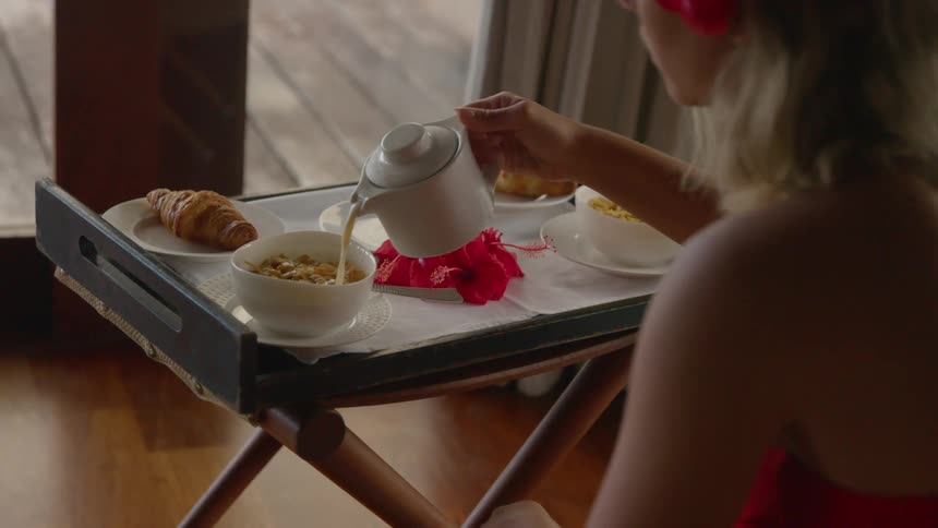 A white woman pours liquid on a bowl of cereal while sitting in a room