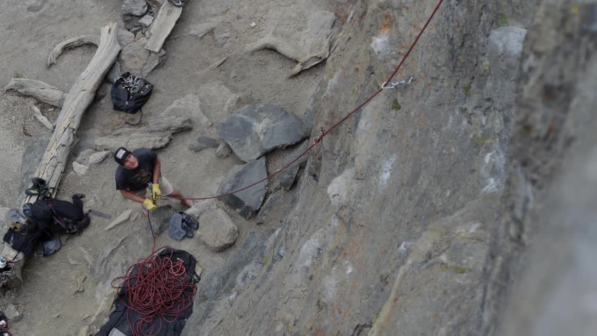 Man surrounded by rock climbing equipment holds a rope attached to a cliff