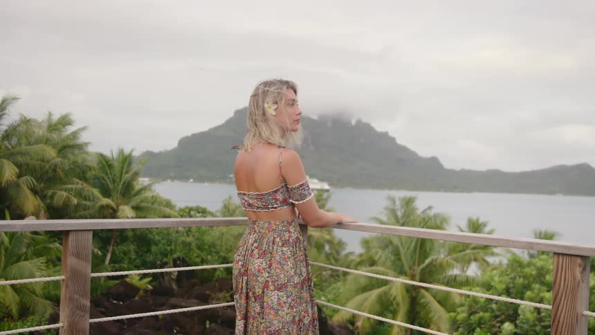 A woman stands on a balcony and looks toward the lake and distant mountain