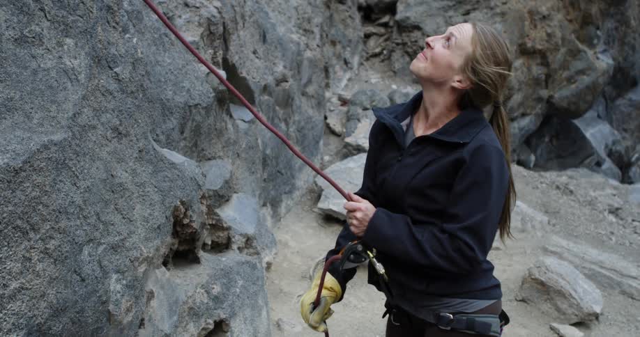 A white woman stands at the bottom of a cliff holding a climbing rope