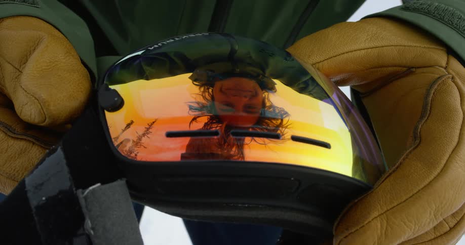 A persons smiling face is colorfully reflected on the ski goggles they are holding in their hands
