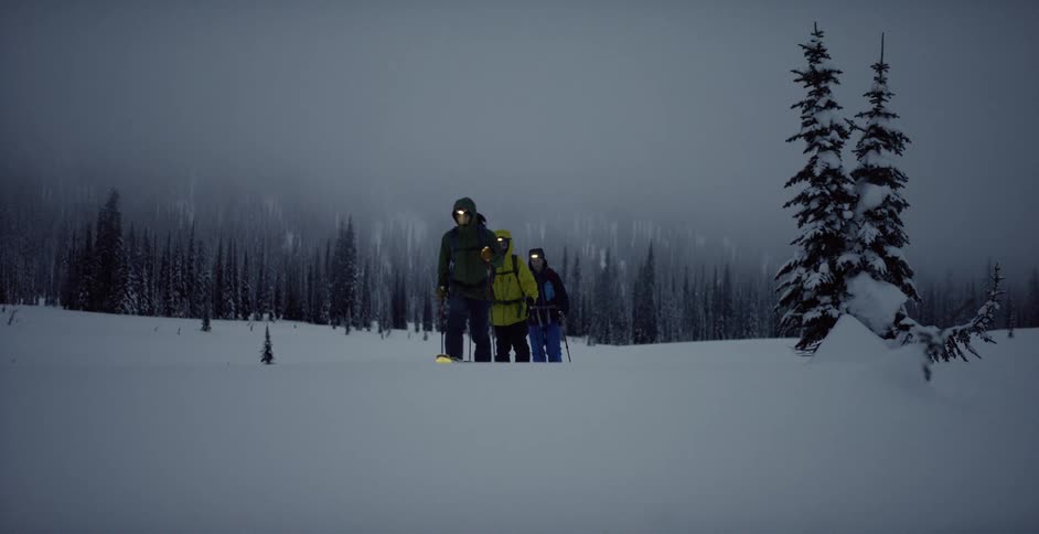 Three people are walking while wearing ski equipment in dim winter light