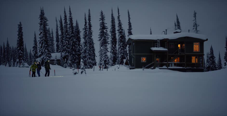 Three people ski away from a house in a rural area in the evening