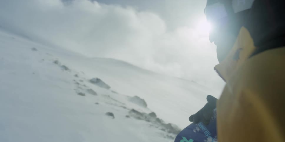 A skier in winter gear stands on a mountain slope and faces a cold wind